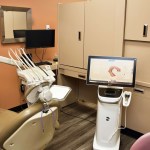 Modern dental treatment room with beige dental chair, computer workstation, and storage cabinets